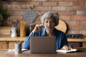 Smart senior grey-haired woman in glasses sit at table at home kitchen look at laptop screen working online. Mature Caucasian 60s female study or watch webinar on computer using internet